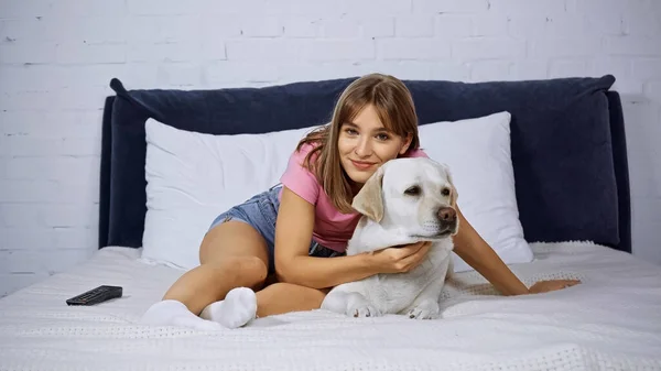 Joyful woman looking at camera near golden retriever and remote controller on bed — Stock Photo