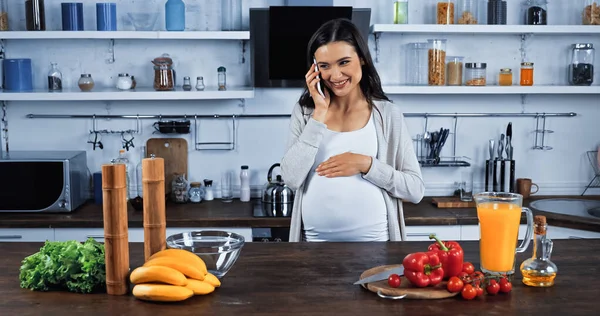 Mulher grávida sorridente falando no smartphone perto de legumes frescos e suco de laranja na cozinha — Fotografia de Stock