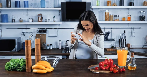 Donna incinta sorridente che scatta foto con smartphone vicino a ingredienti freschi sul tavolo della cucina — Foto stock