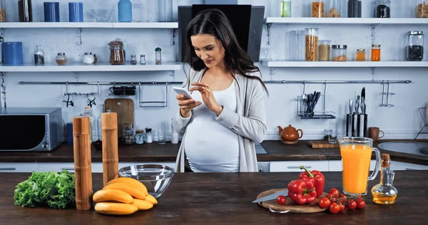 Femme enceinte utilisant un smartphone près des ingrédients bruts dans la cuisine — Photo de stock