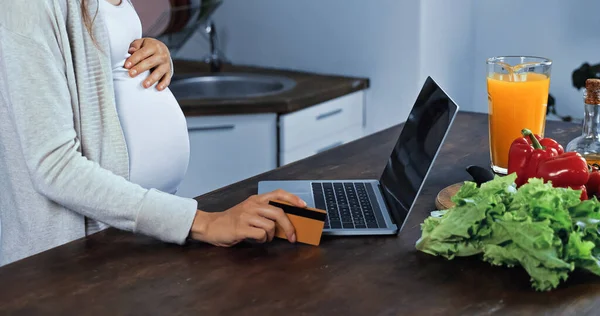 Vista recortada de la mujer embarazada con tarjeta de crédito cerca de la computadora portátil y verduras - foto de stock