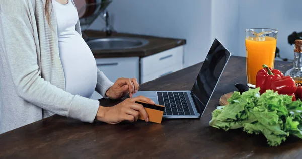 Ausgeschnittene Ansicht einer schwangeren Frau mit Kreditkarte in der Nähe von Laptop und frischem Essen in der Küche — Stockfoto