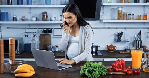 Mujer embarazada usando el ordenador portátil y hablando en el teléfono inteligente cerca de las verduras en la cocina - foto de stock