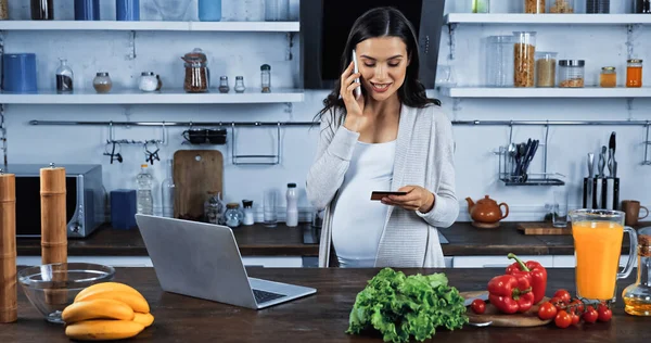 Smiling pregnant woman talking on smartphone while holding credit card near food in kitchen — Stock Photo