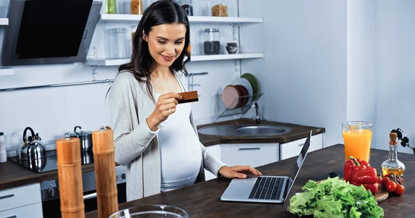 Sourire femme enceinte regardant la carte de crédit près de l'ordinateur portable et des ingrédients sur la table de cuisine — Photo de stock