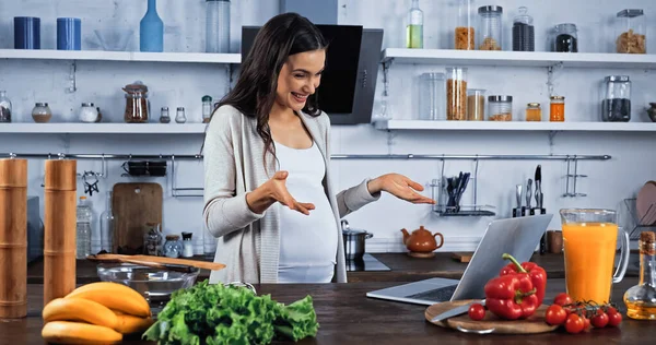 Весела вагітна жінка має відеодзвінок на ноутбук біля їжі на кухні — стокове фото