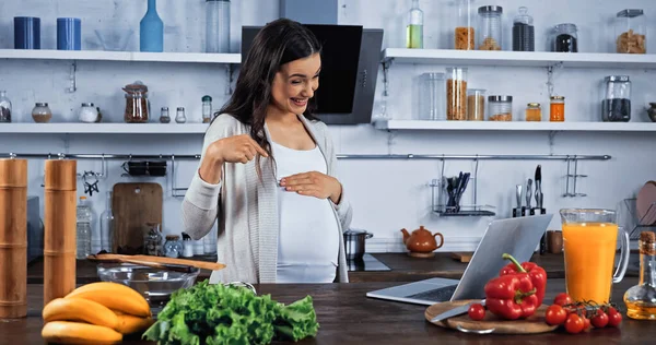 Donna incinta che indica la pancia durante la videochiamata in cucina — Foto stock