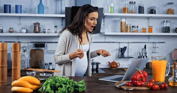 Mujer embarazada emocionada teniendo videollamada en el ordenador portátil cerca de alimentos frescos en la cocina - foto de stock
