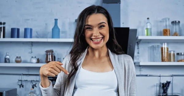 Cheerful pregnant woman pointing at belly in kitchen — Stock Photo