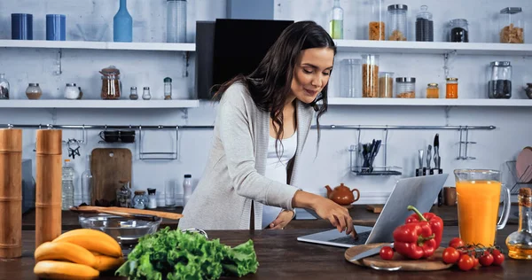 Femme enceinte utilisant un ordinateur portable près des ingrédients frais dans la cuisine — Photo de stock