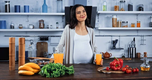 Mujer embarazada sonriente sosteniendo un vaso de jugo de naranja cerca de ingredientes crudos en la cocina - foto de stock