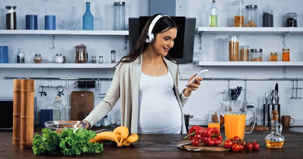 Mujer embarazada sonriente en auriculares con teléfono inteligente cerca de alimentos frescos en la cocina - foto de stock