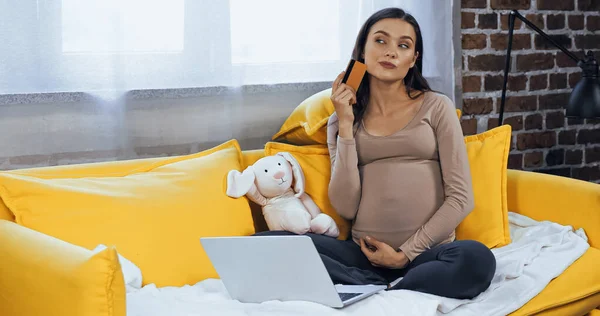 Mujer embarazada pensativa sosteniendo la tarjeta de crédito cerca de la computadora portátil y juguete suave en casa - foto de stock