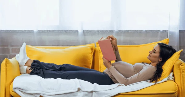 Schwangere liest Buch, während sie zu Hause auf der Couch liegt — Stockfoto