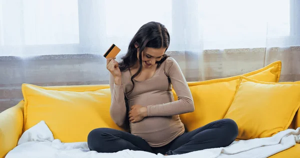 Mujer embarazada sonriente con tarjeta de crédito mirando el vientre en el sofá - foto de stock