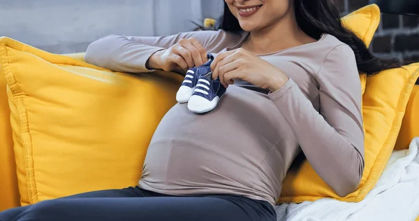Vista recortada de la mujer embarazada sonriendo y sosteniendo botines de bebé cerca del vientre en el sofá - foto de stock
