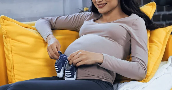 Vista recortada de una mujer embarazada sonriente sosteniendo botines de bebé cerca del vientre - foto de stock