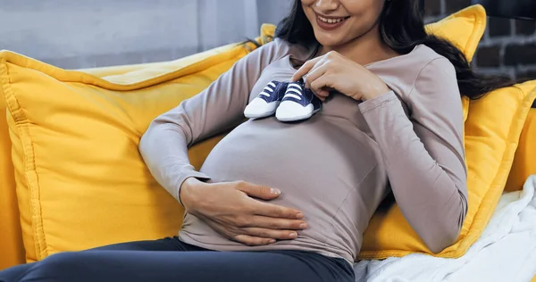 Vista recortada de mujer embarazada sonriente sosteniendo botines de bebé - foto de stock