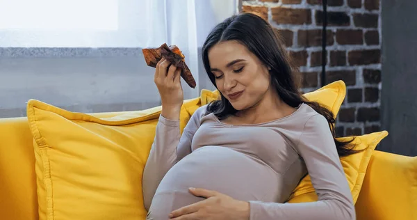 Feliz embarazada comiendo magdalena en el sofá - foto de stock