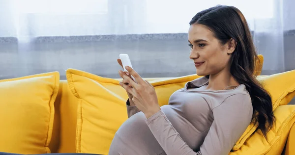 Pregnant woman smiling while using smartphone on sofa in living room — Stock Photo