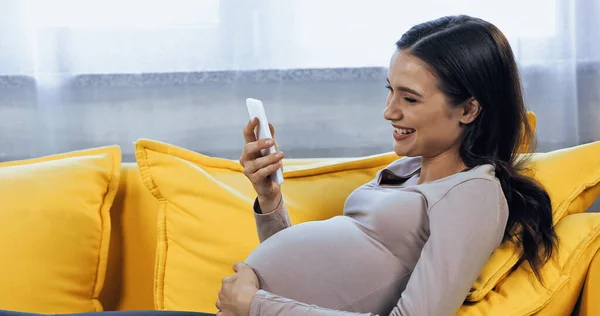 Улыбающаяся беременная женщина с помощью смартфона, лежа на диване — стоковое фото