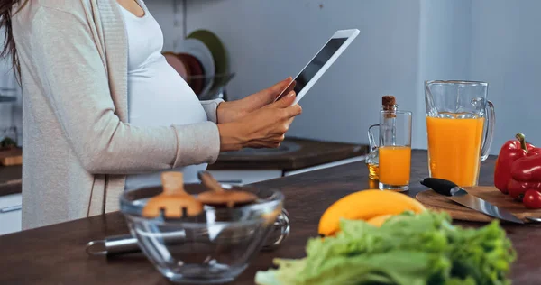 Vista cortada da mulher grávida segurando comprimido digital perto de alimentos frescos — Fotografia de Stock