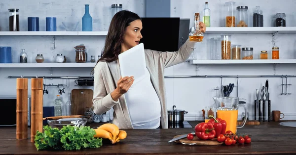 Mujer embarazada con tableta digital mirando el aceite cerca de la comida en la mesa de la cocina - foto de stock