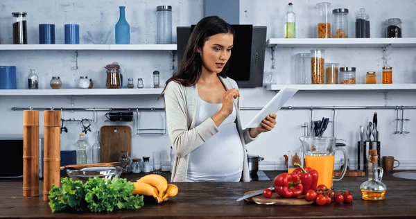 Беременная женщина с цифровым планшетом рядом с едой на кухонном столе — стоковое фото