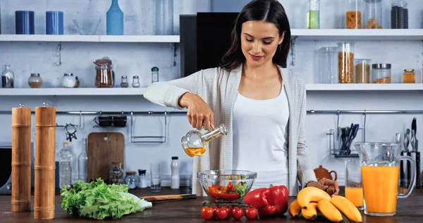 Брюнетка наливает масло в салат рядом со свежими ингредиентами на кухне — стоковое фото
