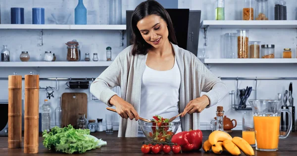 Улыбающаяся женщина смешивает свежий салат рядом с апельсиновым соком на кухонном столе — стоковое фото