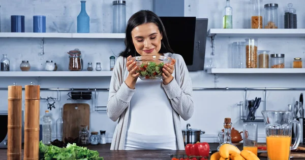 Smiling woman holding bowl with fresh salad near vegetables and orange juice — Stock Photo