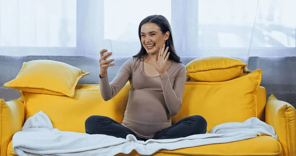 Mujer embarazada saludando de la mano durante el chat de vídeo en el teléfono inteligente en casa - foto de stock