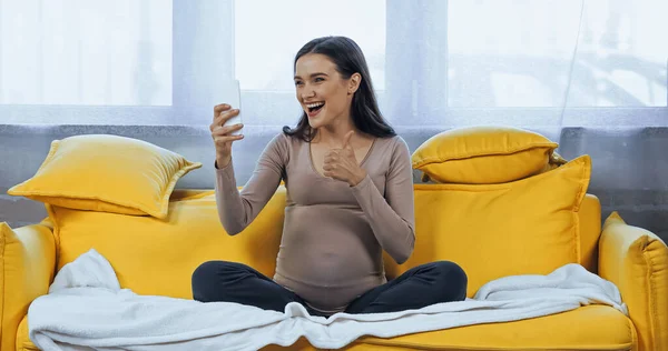 Mujer embarazada alegre mostrando como durante la videollamada en el teléfono inteligente - foto de stock