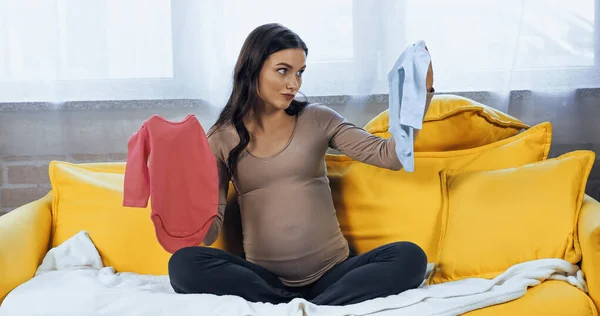Беременная женщина смотрит на детский костюм на диване — стоковое фото