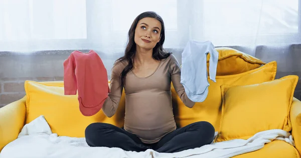 Pensive pregnant woman holding baby bodysuits in living room — Stock Photo