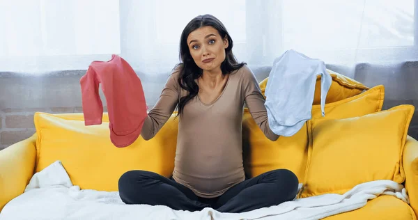 Mulher grávida confusa segurando roupas de bebê no sofá — Fotografia de Stock