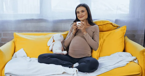 Dreamy pregnant woman with cup sitting near soft toy — Stock Photo