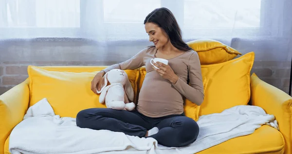 Беременная женщина с чашкой смотрит на мягкую игрушку, сидя на желтом диване — стоковое фото