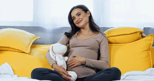 Pregnant woman with soft toy smiling away on yellow couch — Stock Photo