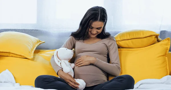Беременная женщина улыбается, обнимая живот и мягкую игрушку — стоковое фото