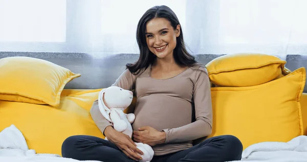 Pregnant woman holding soft toy and smiling at camera on couch — Stock Photo