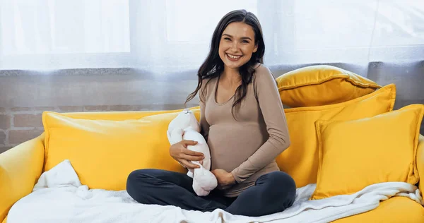 Schwangere Brünette lächelt in die Kamera und hält Stofftier auf Couch — Stockfoto