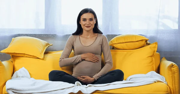 Schwangere umarmt Bauch und lächelt auf Sofa in die Kamera — Stockfoto
