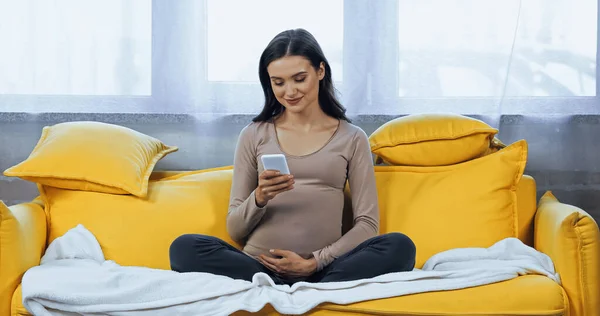 Pregnant brunette woman smiling and using smartphone on couch — Stock Photo