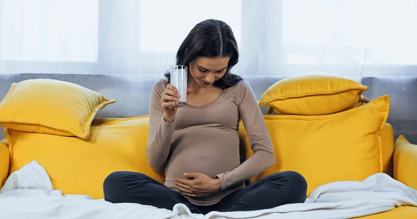 Pregnant woman holding glass of fresh milk and looking at belly on sofa — Stock Photo