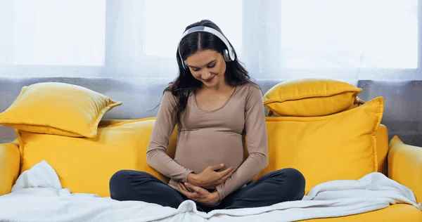 Lächelnde Schwangere mit Kopfhörer umarmt Bauch im Wohnzimmer — Stockfoto