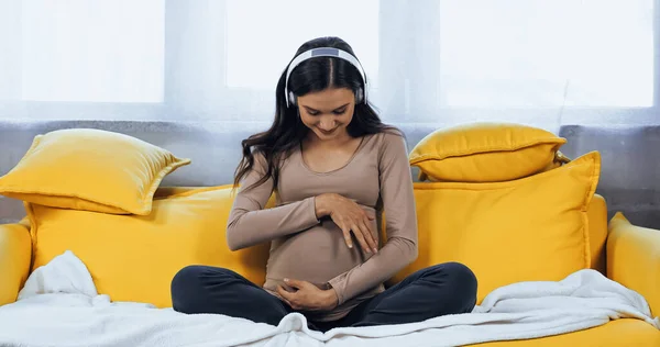 Pregnant woman in headphones looking at belly on couch — Stock Photo