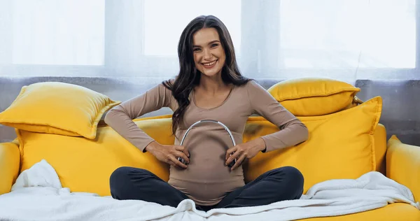 Mulher grávida alegre segurando fones de ouvido perto da barriga — Fotografia de Stock