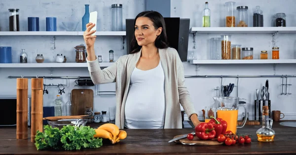 Mujer embarazada tomando selfie cerca de ingredientes crudos en la mesa de la cocina - foto de stock