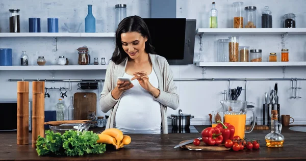 Donna incinta sorridente utilizzando smartphone vicino al cibo crudo sul tavolo della cucina — Foto stock